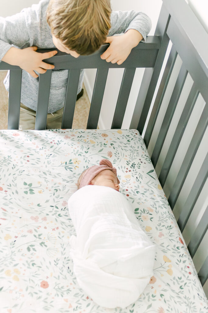 little boy looks into crib where newborn baby sister is sleeping, dayton ohio photographer takes pictures