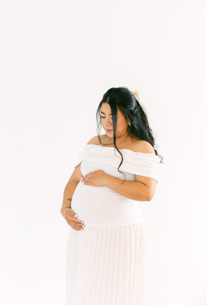 Pregnant mother in a flowing white dress gently holding her baby bump during a timeless studio maternity session by a Cincinnati Maternity Photographer.