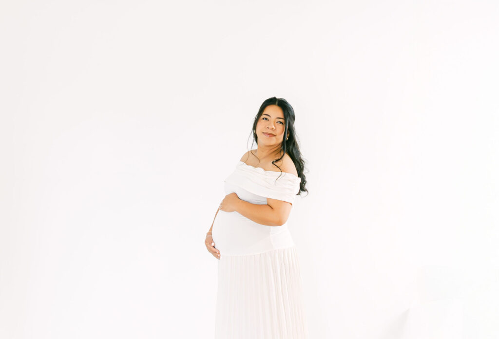 Pregnant mother in a flowing white dress gently holding her baby bump during a timeless studio maternity session by a Cincinnati Maternity Photographer.