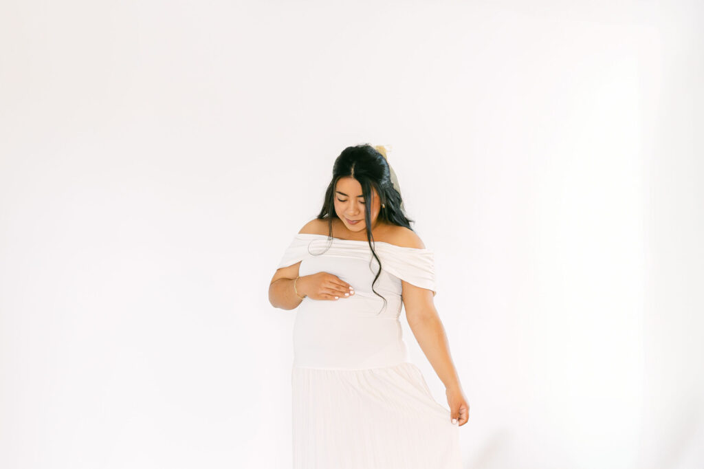 Pregnant mother in a flowing white dress gently holding her baby bump during a timeless studio maternity session by a Cincinnati Maternity Photographer.