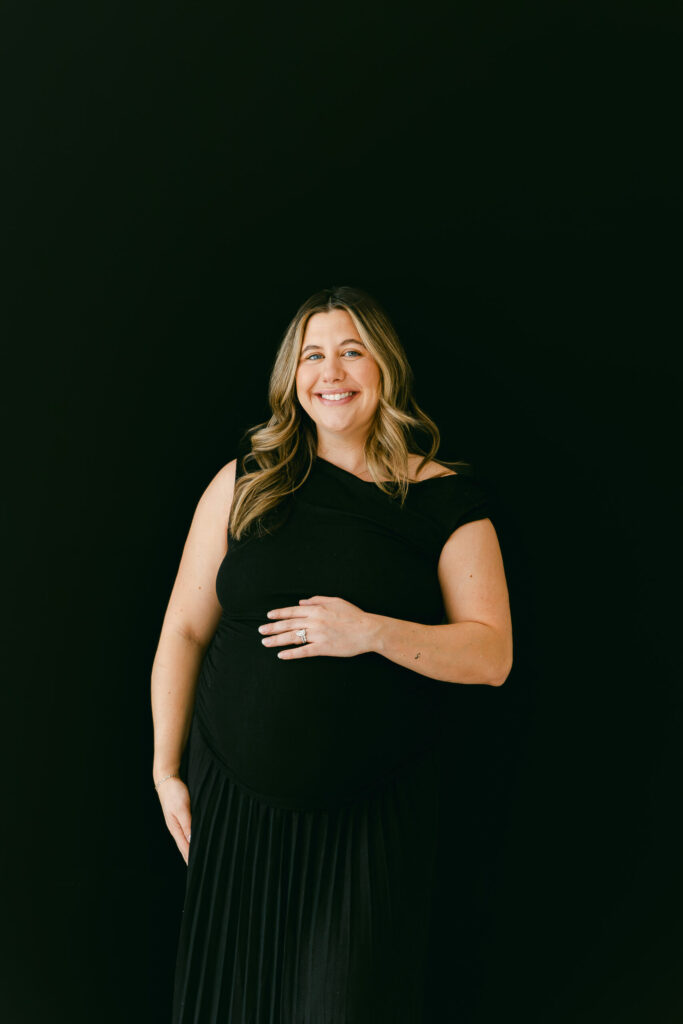 mom smiling at camera with hand on the bump with black backdrop and back maternity dress